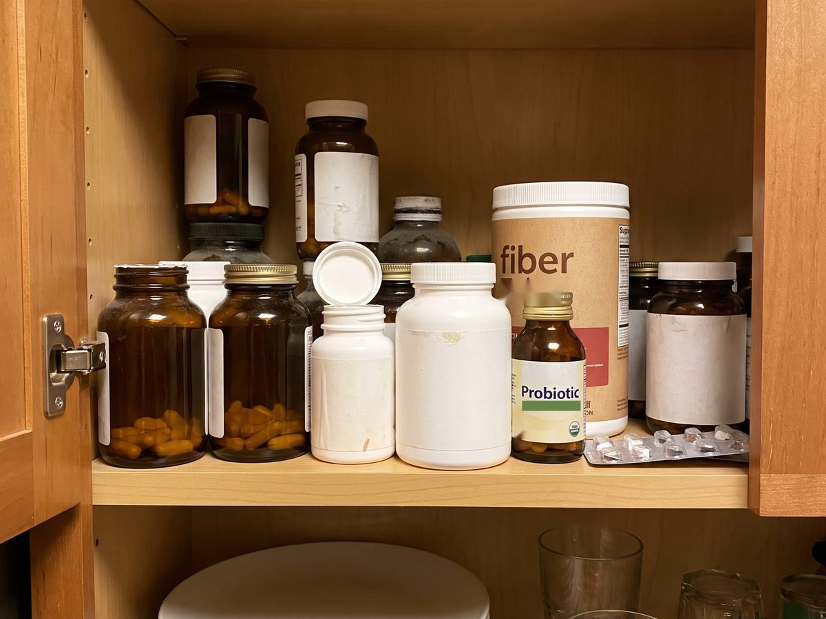 A kitchen cabinet shelf crowded with half-used supplement bottles